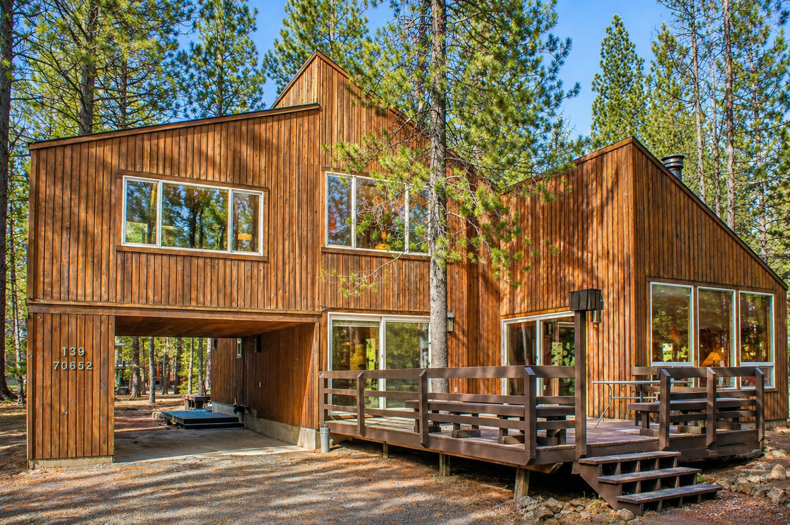 A stunning modern cedar cabin nestled among towering pine trees, featuring a covered carport and wraparound deck.