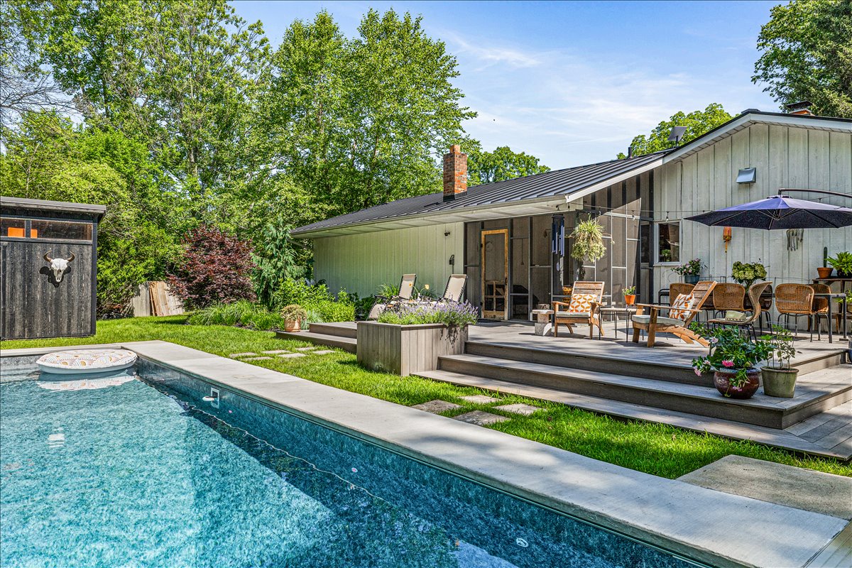 Modern retreat featuring a sparkling pool, elevated deck with lounge seating, and lush landscaped grounds surrounded by mature trees.