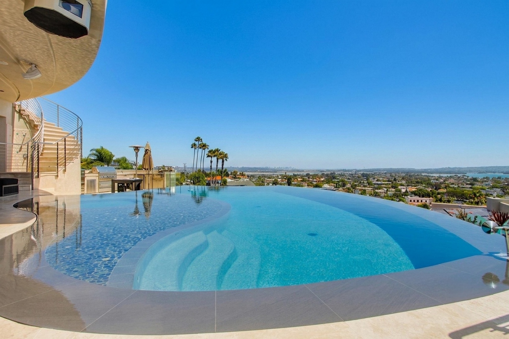 Stunning infinity pool overlooking coastal neighborhoods and city skyline, with palm trees and outdoor seating in a resort-style hillside setting.