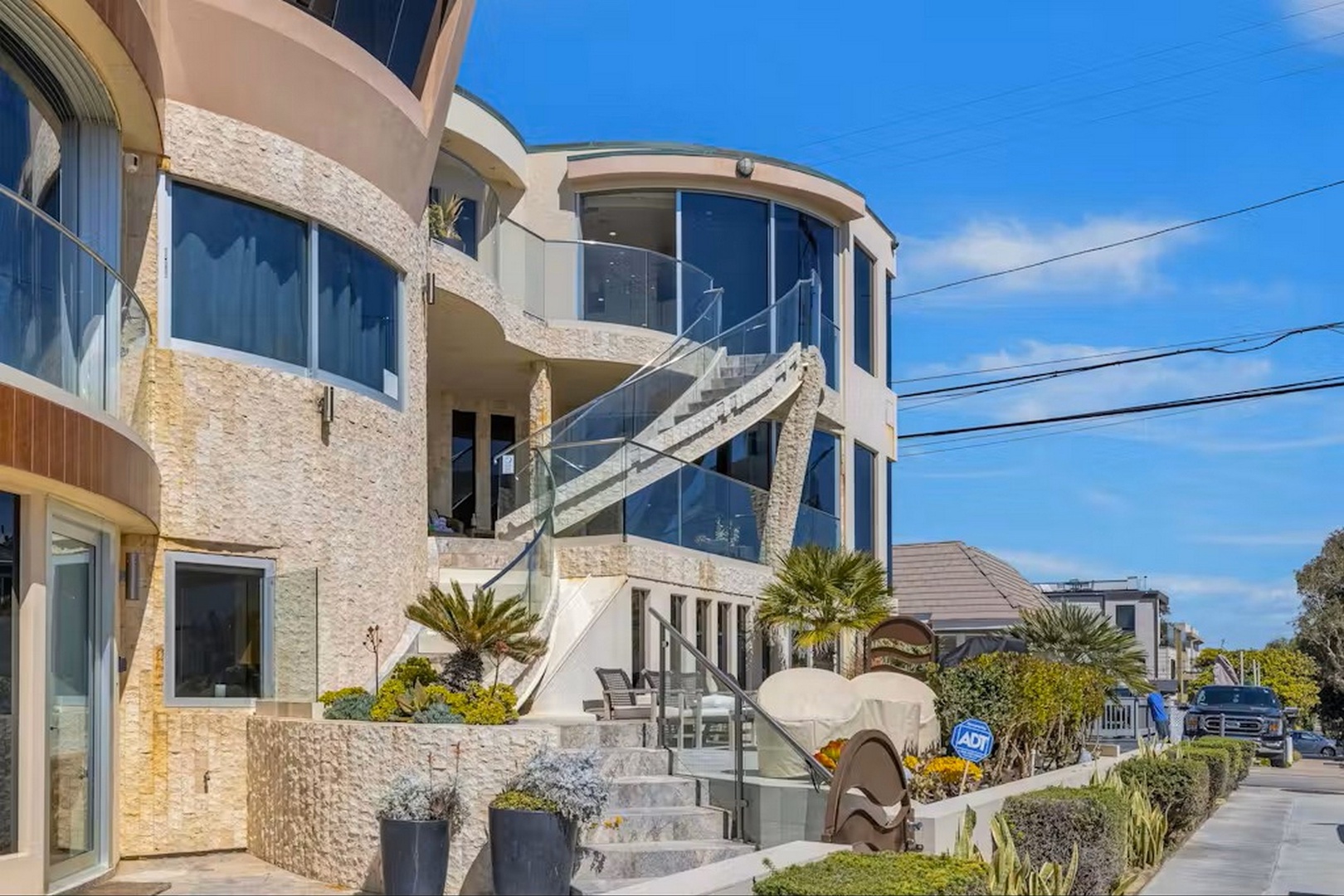 A striking modern beachfront property featuring curved architecture with expansive floor-to-ceiling windows, glass-railed balconies, and tropical landscaping positioned steps from the ocean.