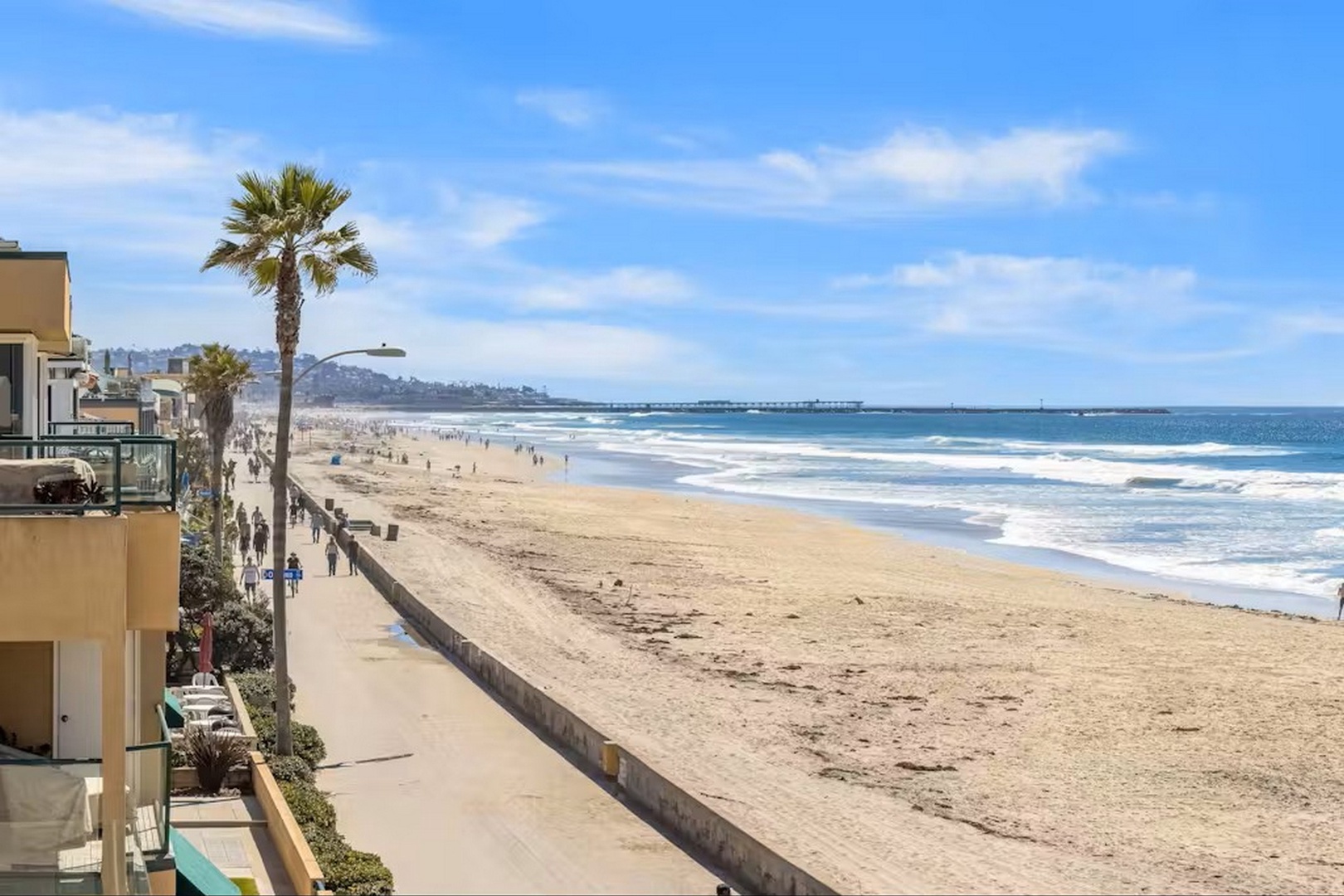 Beachfront property situated along pristine sandy shores with palm trees, boardwalk access, and sweeping ocean views extending to the pier.