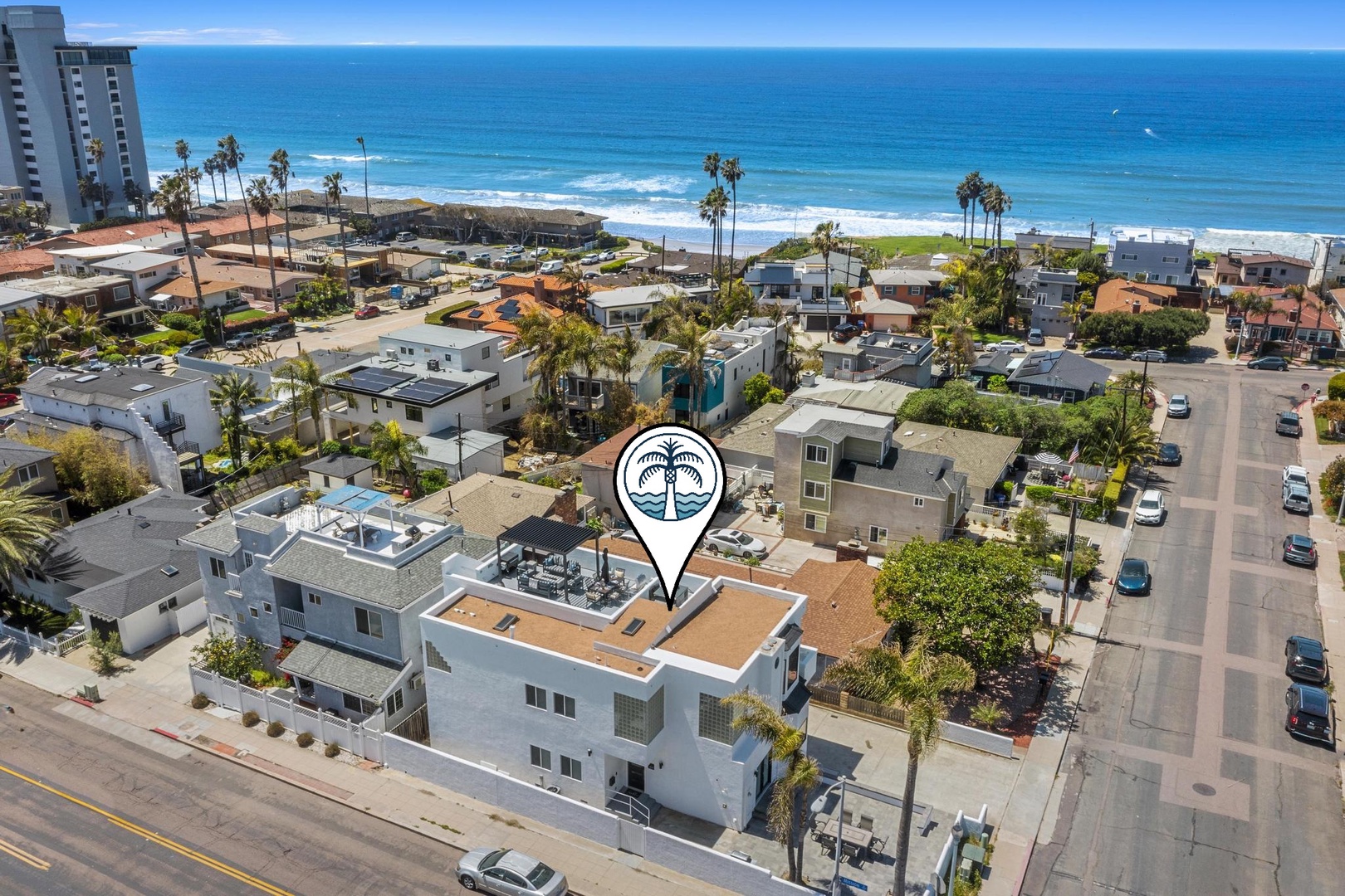 Aerial view of a coastal vacation rental property steps from the ocean, with palm trees dotting the beachfront neighborhood and panoramic sea views stretching to the horizon.