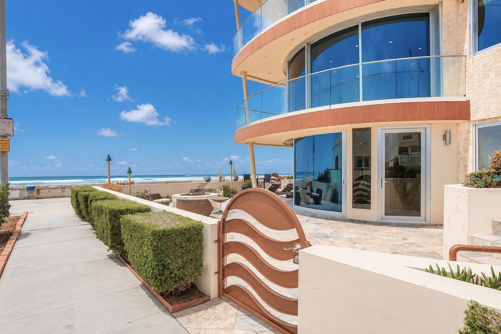 Modern beachfront property featuring curved glass architecture and direct beach access. Manicured hedges and a decorative gate frame the entrance to this oceanside vacation rental.