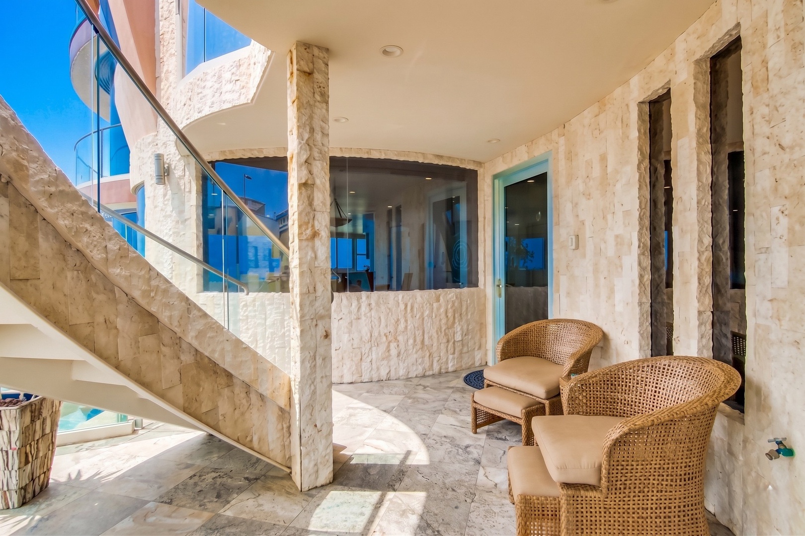 Relax in your private covered terrace, where elegant stone columns frame cozy wicker seating and glass doors open to reveal sleek indoor spaces beyond.