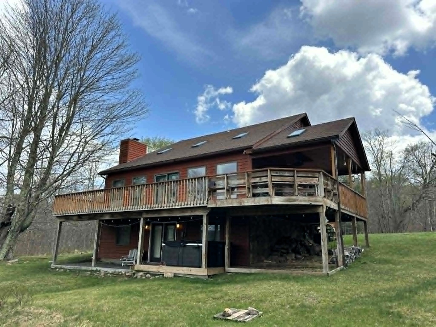 A rustic two-story retreat featuring wraparound deck and natural stone accents, nestled among mature trees on expansive green grounds.