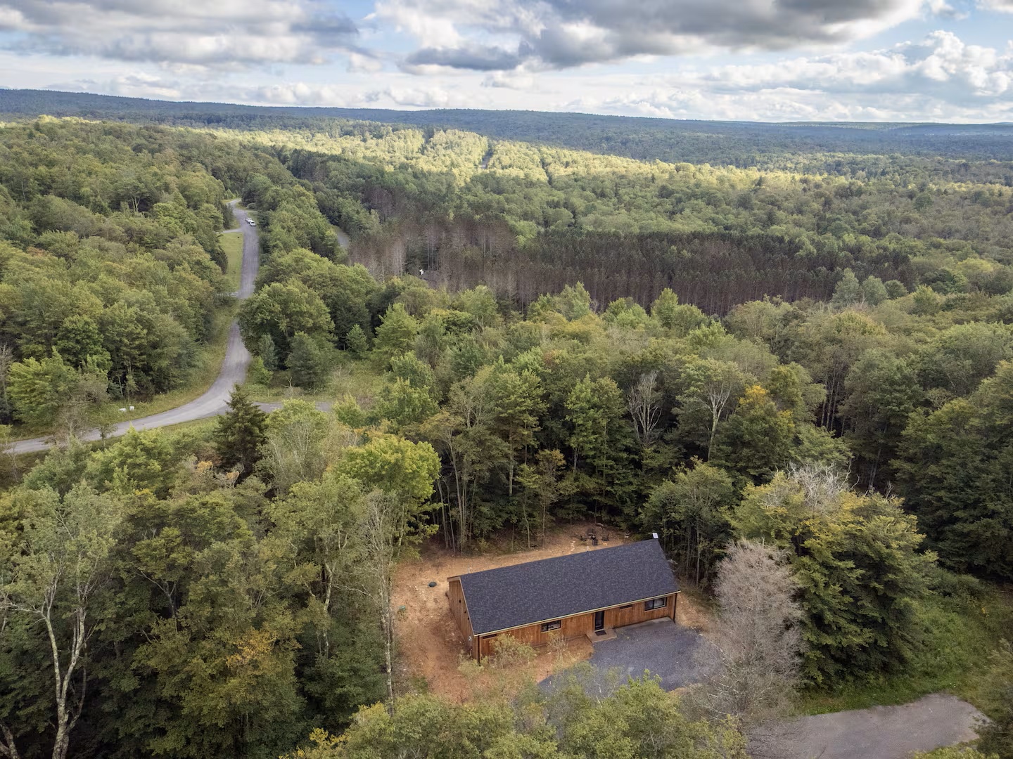 An aerial view captures the property nestled within lush forest, offering a peaceful, secluded retreat surrounded by towering trees and rolling hills under a dramatic sky.