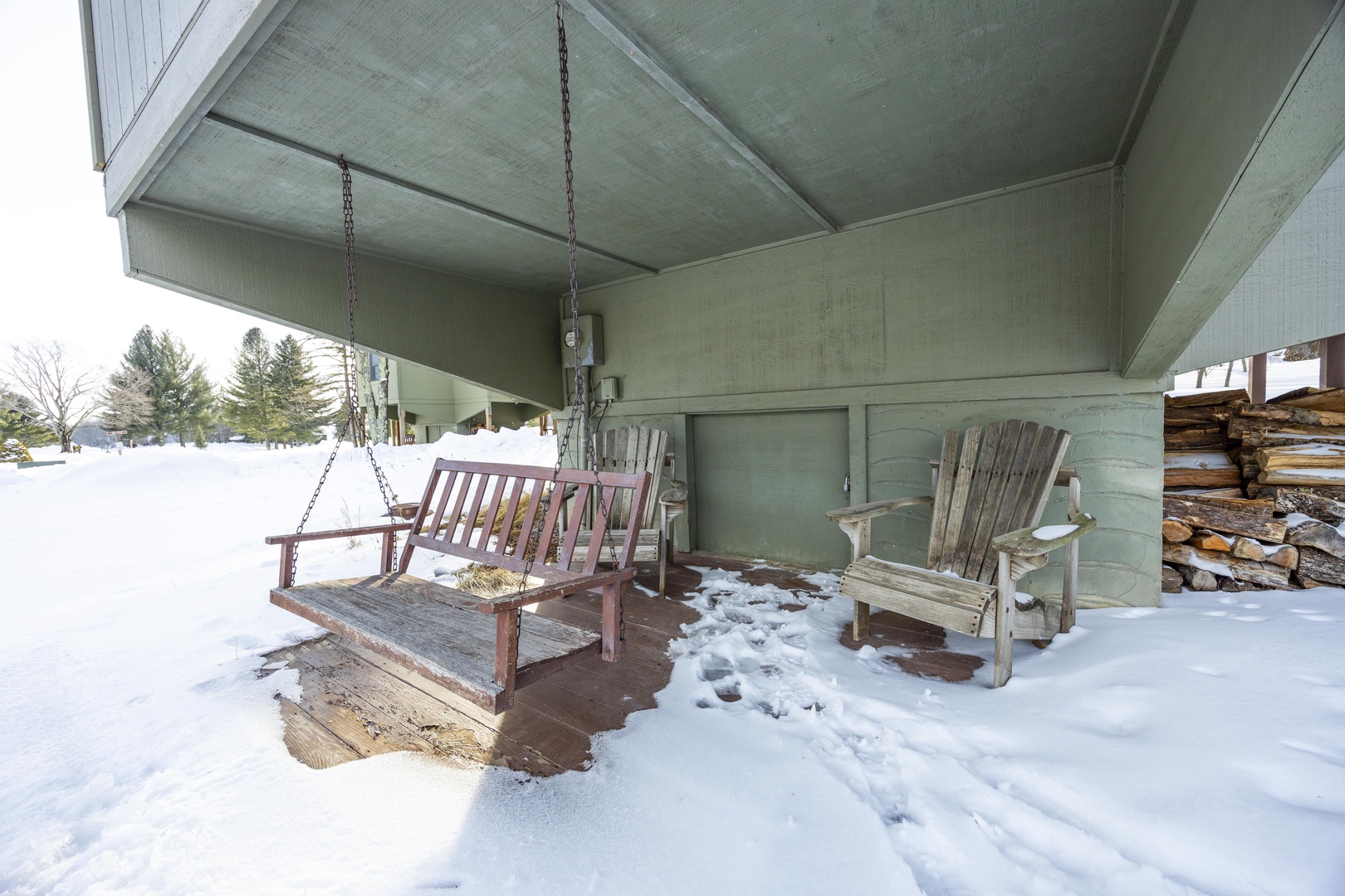 Covered porch with a hanging swing and wooden Adirondack chairs surrounded by snow-draped landscape—a peaceful winter retreat with stacked firewood nearby for cozy evenings.