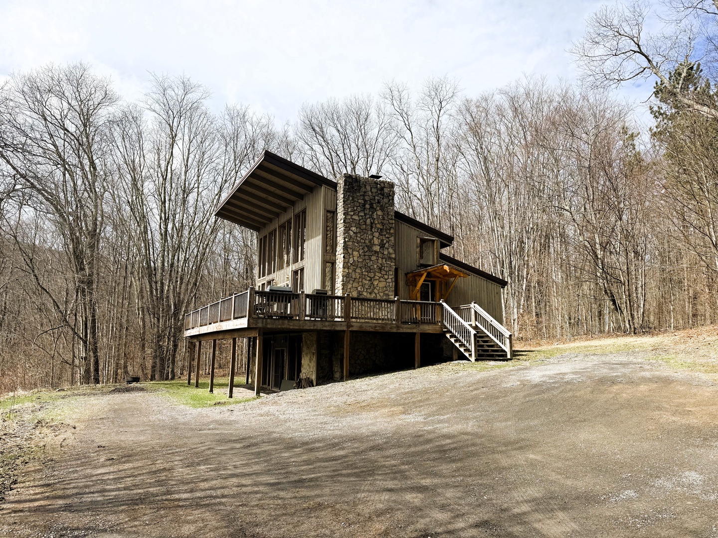 Modern cabin with stone chimney nestled among bare trees, featuring elevated deck and forest access.