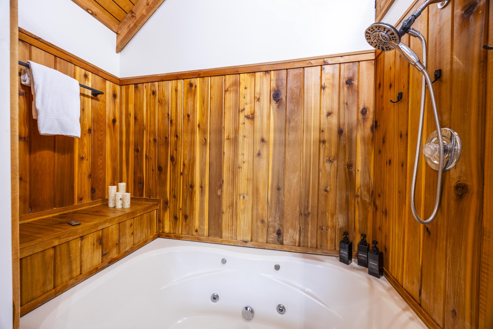 Soak in the warmth of this rustic-luxe bathroom with a jetted tub and handheld shower, surrounded by glowing wood paneling and vaulted beams above.