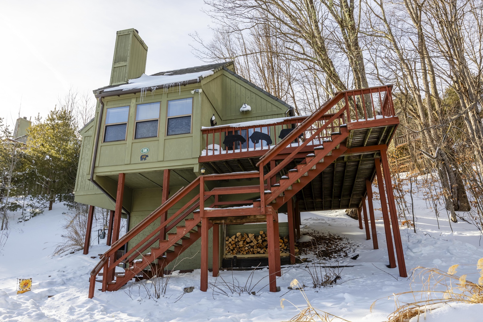 A cozy green cabin nestled in a snowy woodland setting, accessed by a two-level wooden deck with stairs. Firewood stacked beneath, surrounded by bare winter trees.