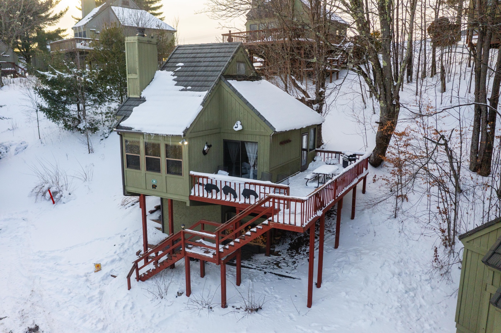 Charming green cottage nestled among snow-covered trees with a welcoming elevated deck, offering a peaceful winter escape in a wooded hillside setting.