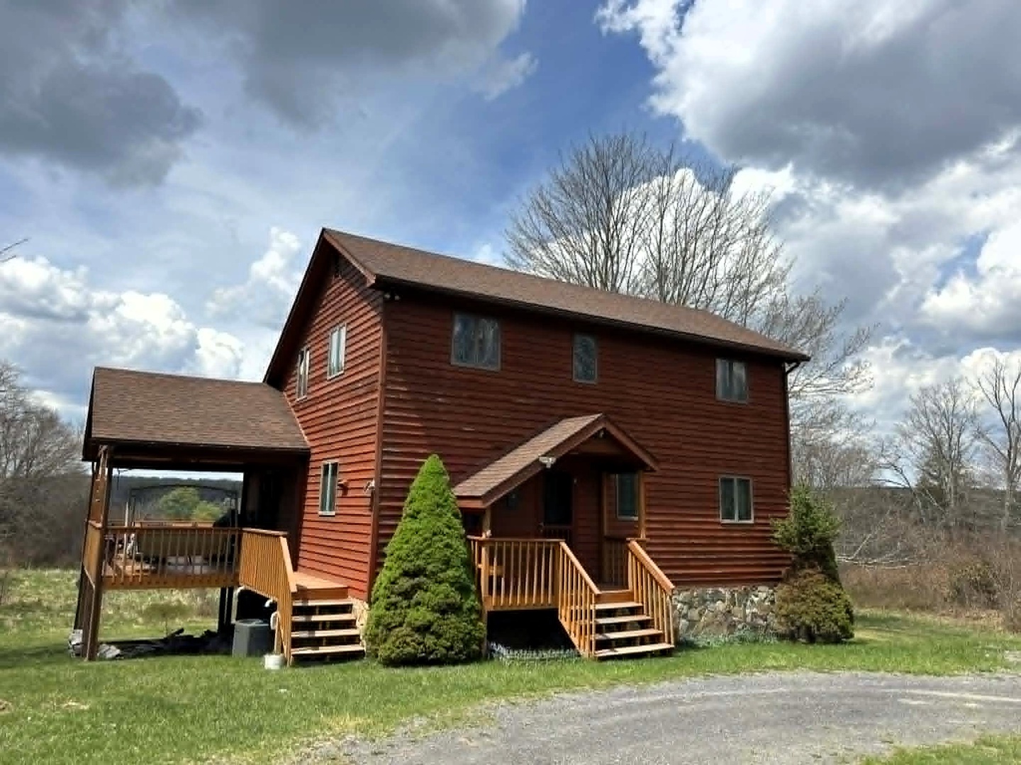 Charming log cabin with covered deck surrounded by open fields and mature trees under a dramatic sky.