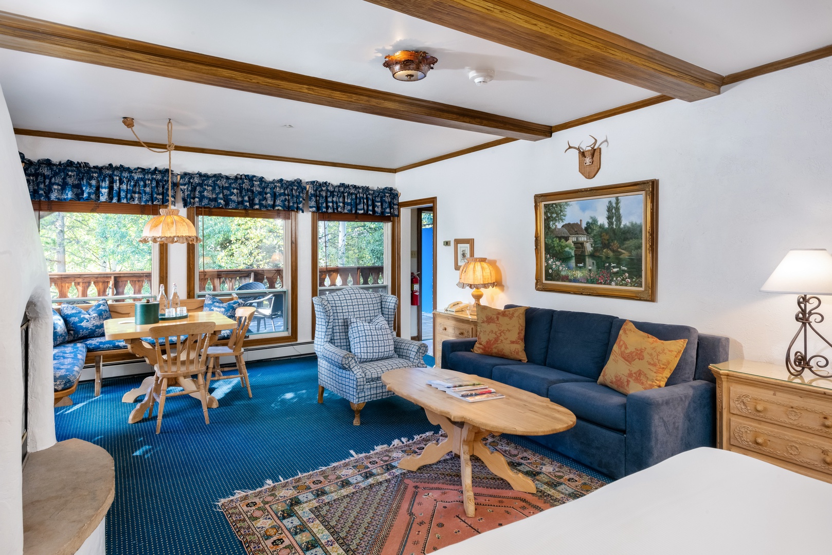 Cozy alpine retreat featuring rustic wooden beams and warm blue tones. Unwind on the plush sofa or enjoy meals at the dining nook overlooking the forested balcony.