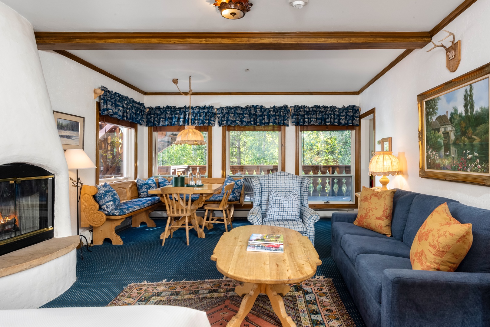 Unwind in your cozy alpine living room with a crackling fireplace, built-in dining nook, and mountain-fresh views through sunny windows that frame the lush greenery just outside.