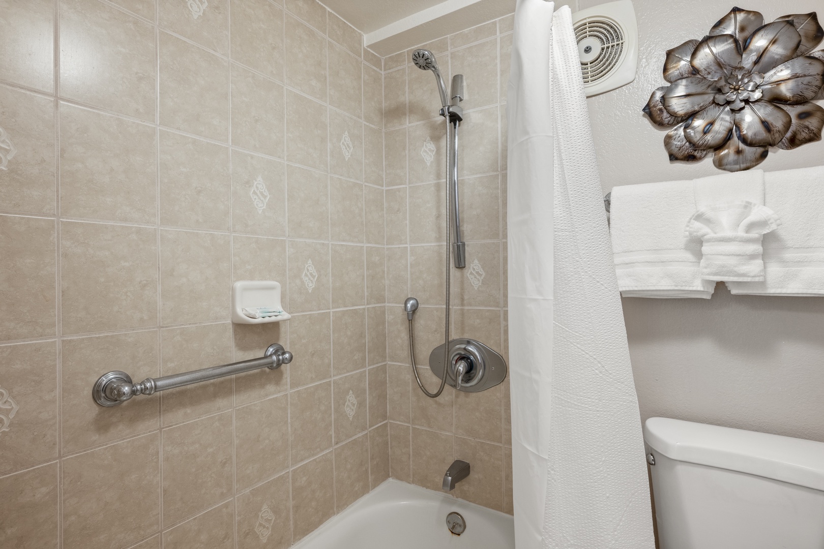 Your bathroom features elegant beige tiles, a convenient grab bar for safety, and plush towels waiting for you. The handheld showerhead adds a thoughtful touch to your daily routine.