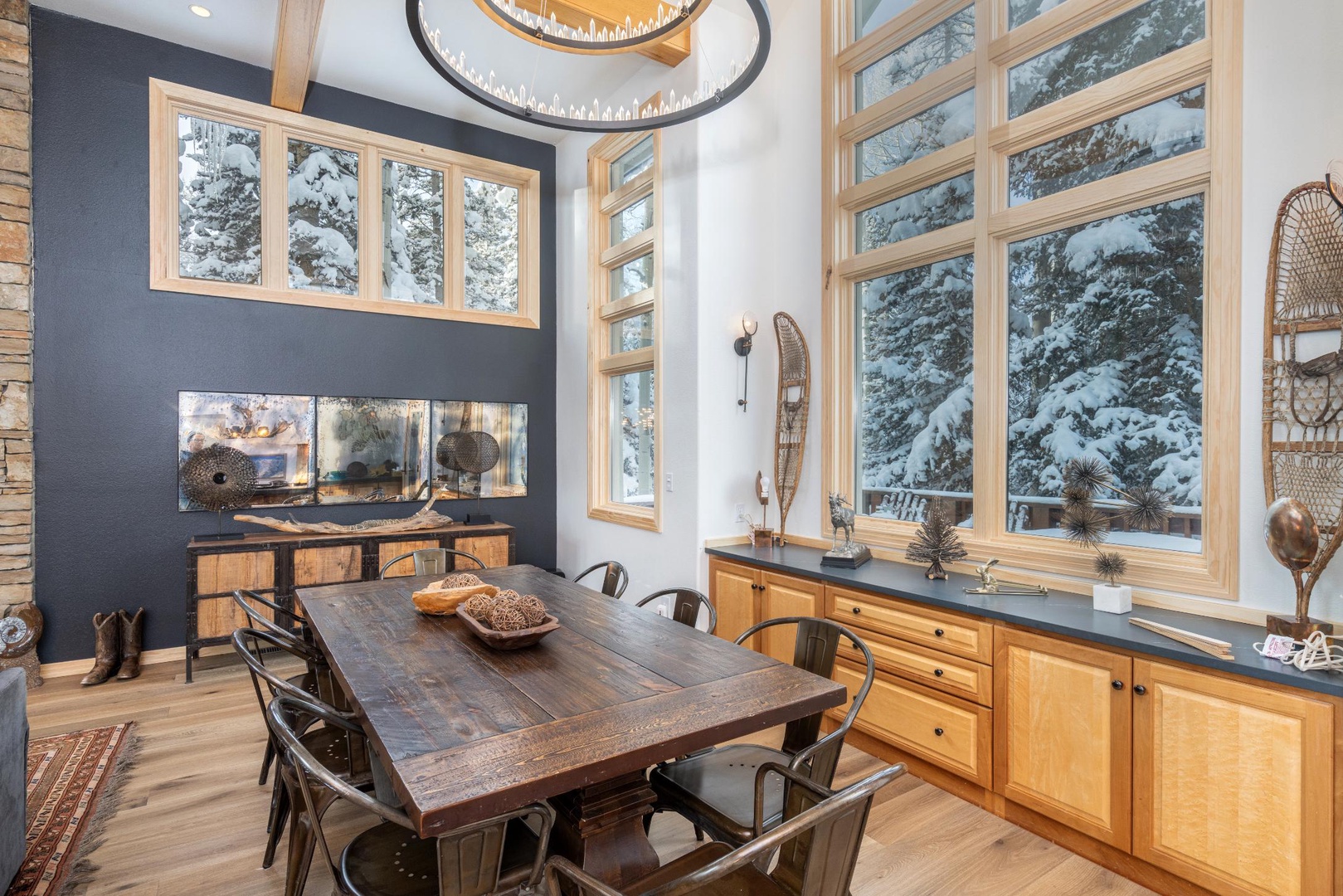 Gather around your rustic dining table with friends and family while enjoying panoramic winter forest views through floor-to-ceiling windows and cozy fireplace ambiance.