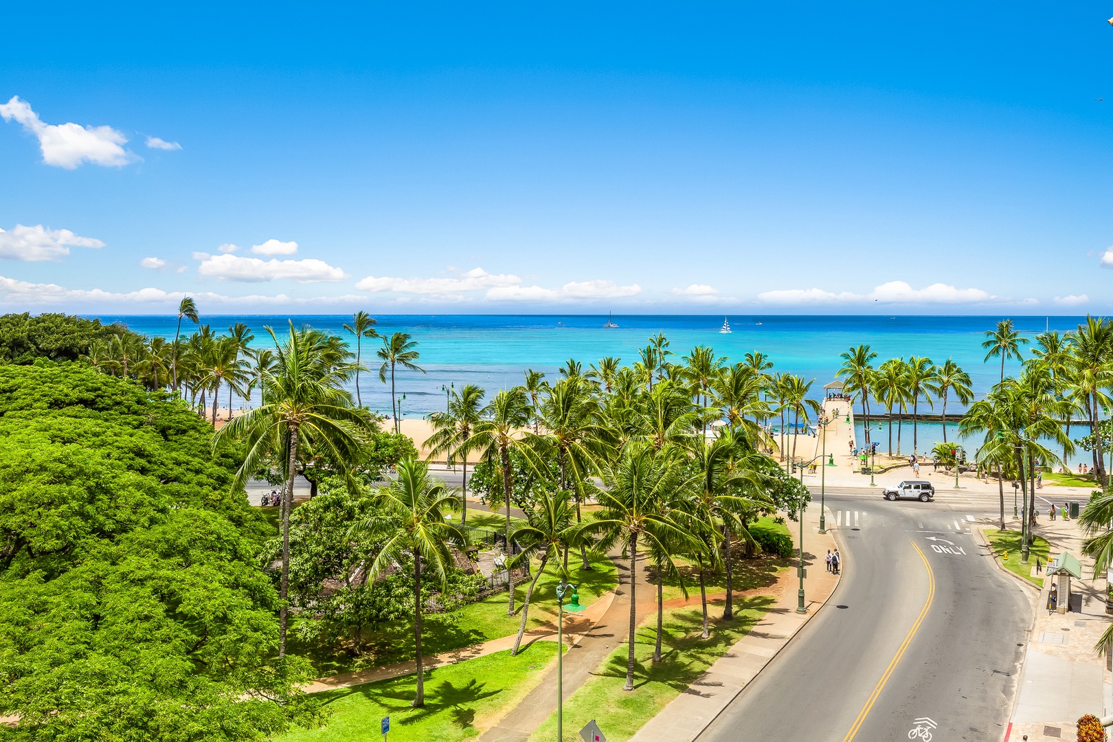 A scenic coastal property location features palm-lined streets leading to a pristine beachfront with turquoise waters and distant sailboats under clear blue skies.