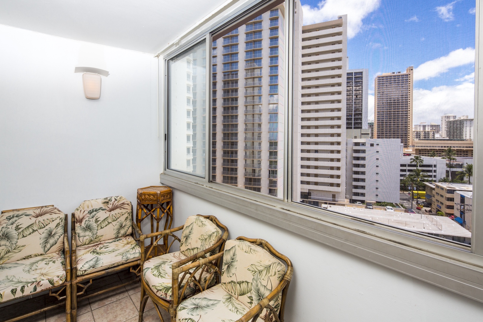 An enclosed balcony with high-rise city views features tropical-print seating, offering a bright and breezy spot to relax while overlooking the urban skyline and distant ocean.