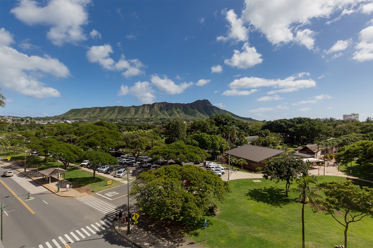 Iconic Diamond Head crater rises majestically above lush tree-lined streets and manicured green spaces, framing this tropical Waikiki location with stunning natural beauty.