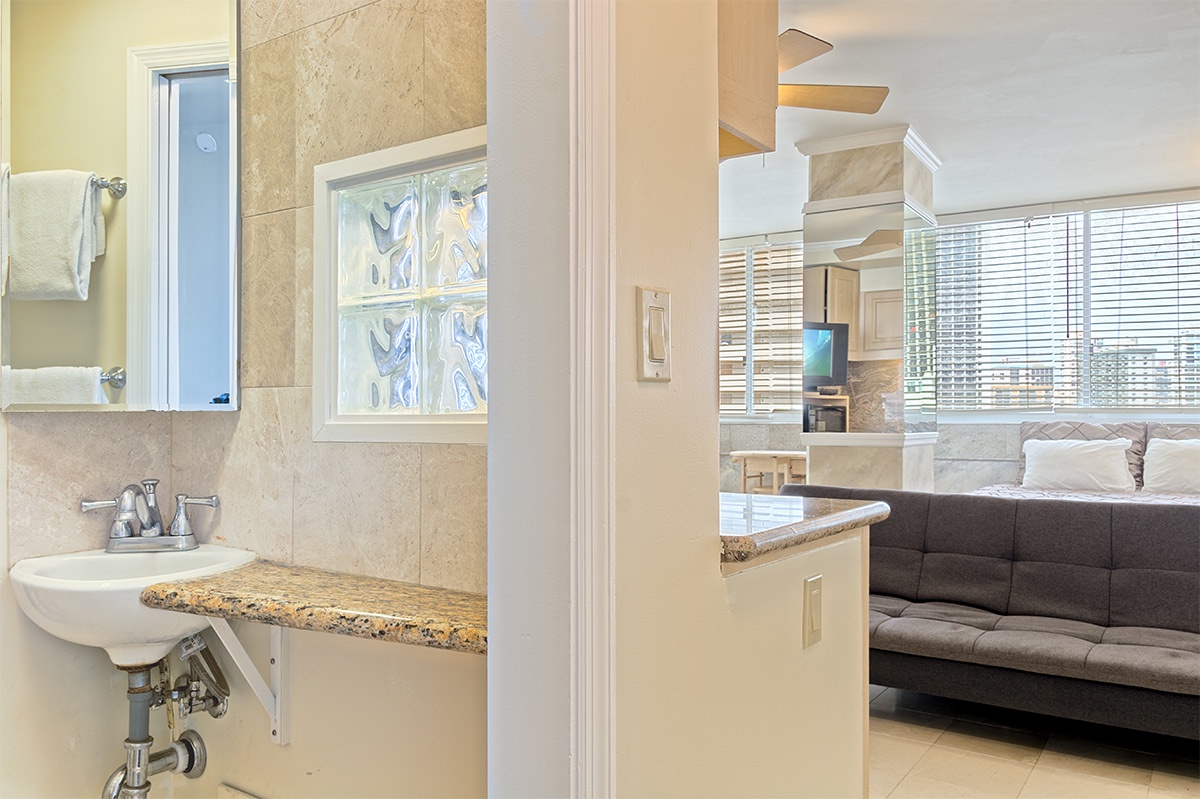 Your private bathroom features warm tones, natural light from a decorative glass block window, and a granite vanity. Beyond, your open living space awaits with modern furnishings.
