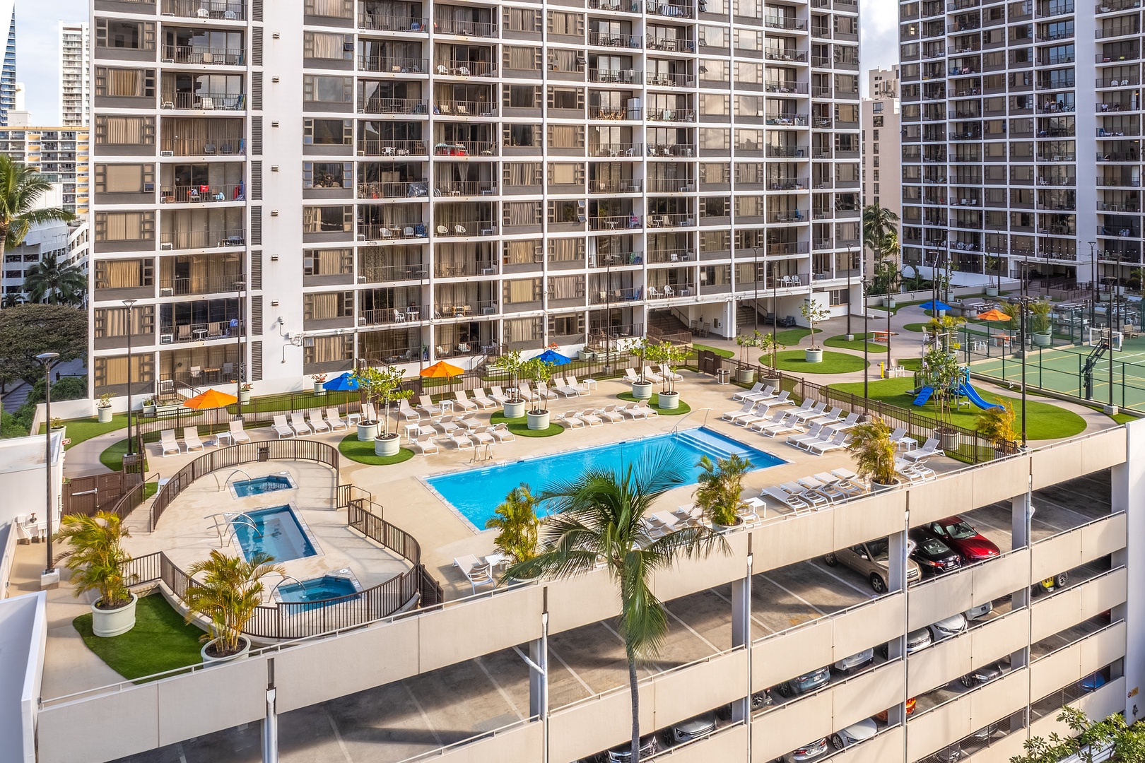 An elevated amenity deck features a sparkling pool, hot tubs, and surrounding green spaces with lounge seating set against a backdrop of high-rise buildings.