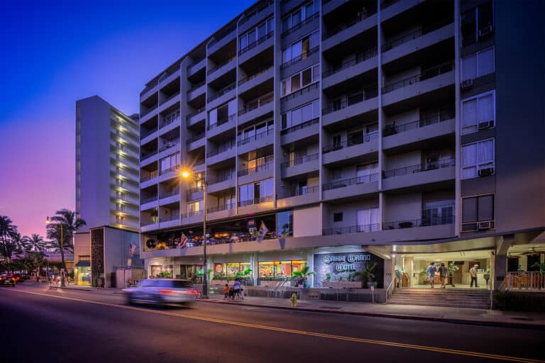 The property's vibrant street-level entrance illuminates the urban landscape at dusk, with palm trees lining the sidewalk and a welcoming lobby that invites guests to explore the area's tropical charm.