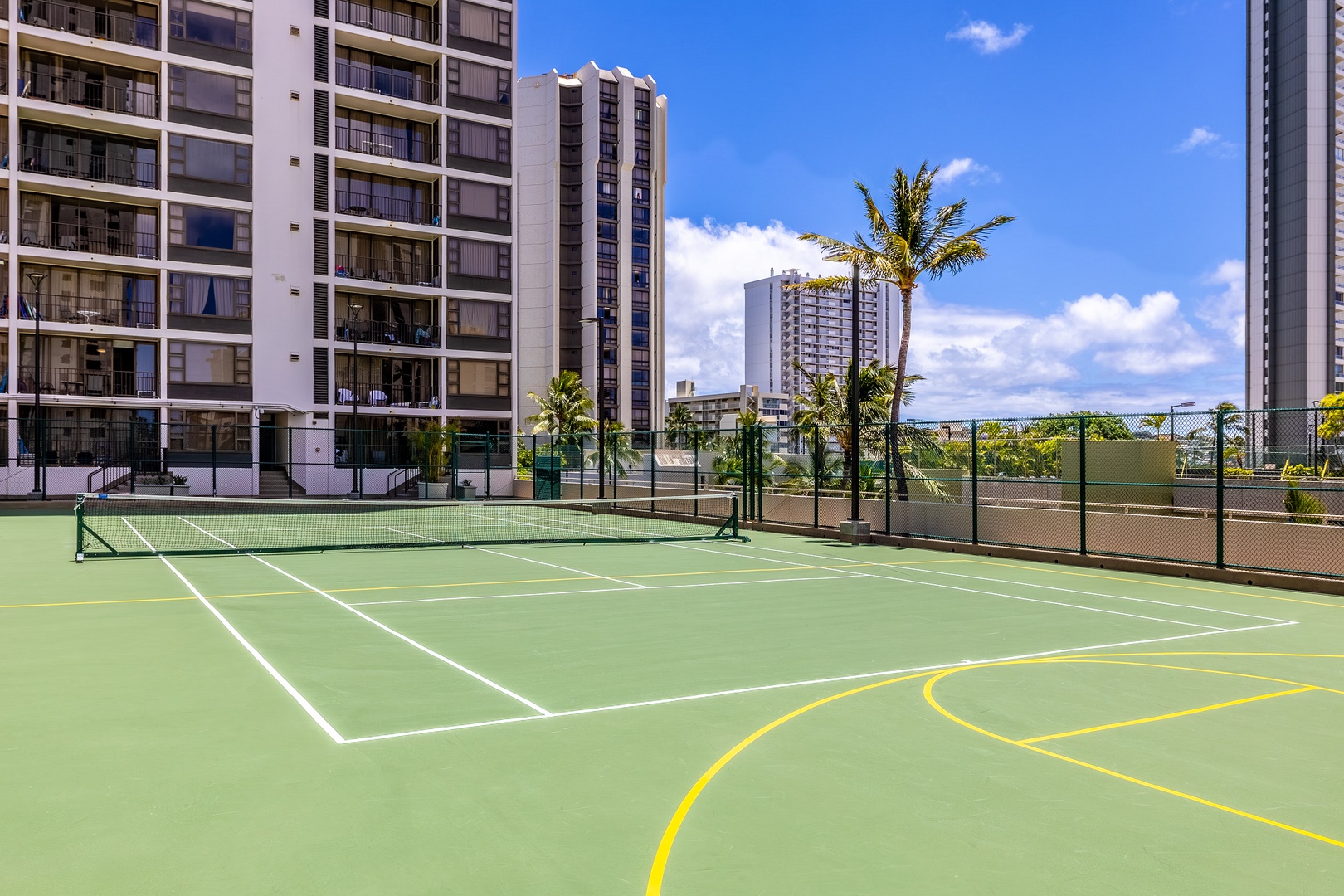 A rooftop tennis court surrounded by palm trees and high-rise buildings, located in the property area.