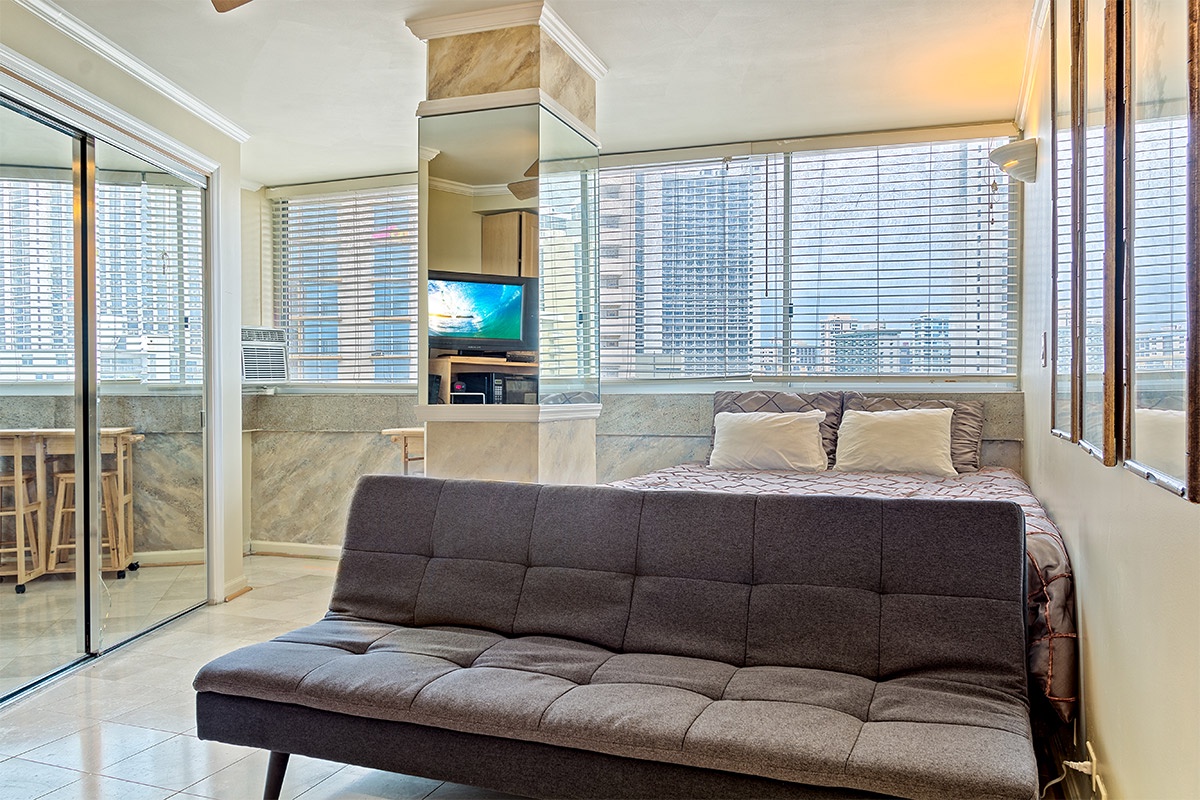 Your bright, modern studio features floor-to-ceiling windows with city views, a plush sofa, comfortable sleeping area, and seamless balcony access—perfect for urban relaxation.