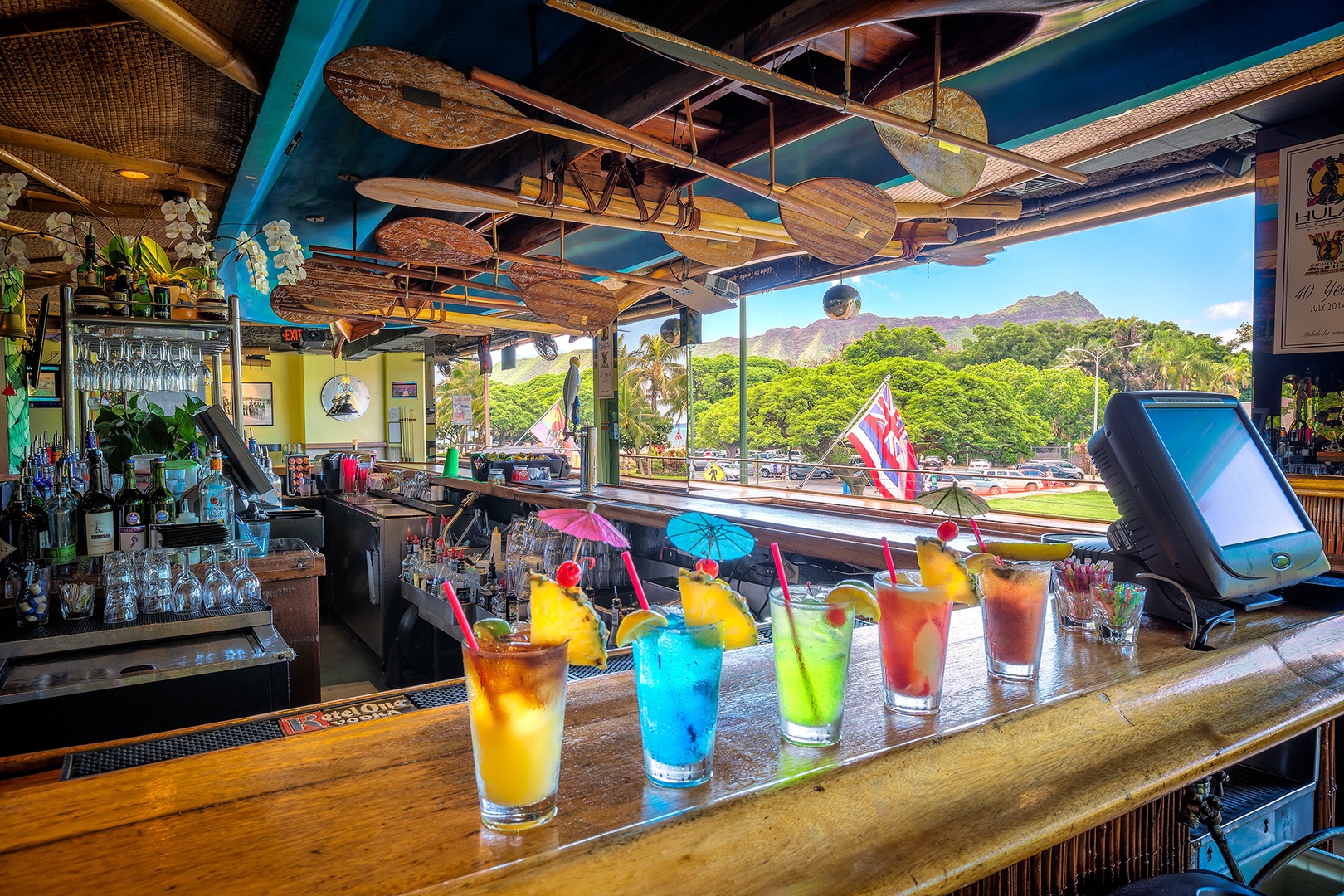 A vibrant tropical bar featuring colorful cocktails, vintage décor, and a scenic mountain backdrop visible through large open windows.