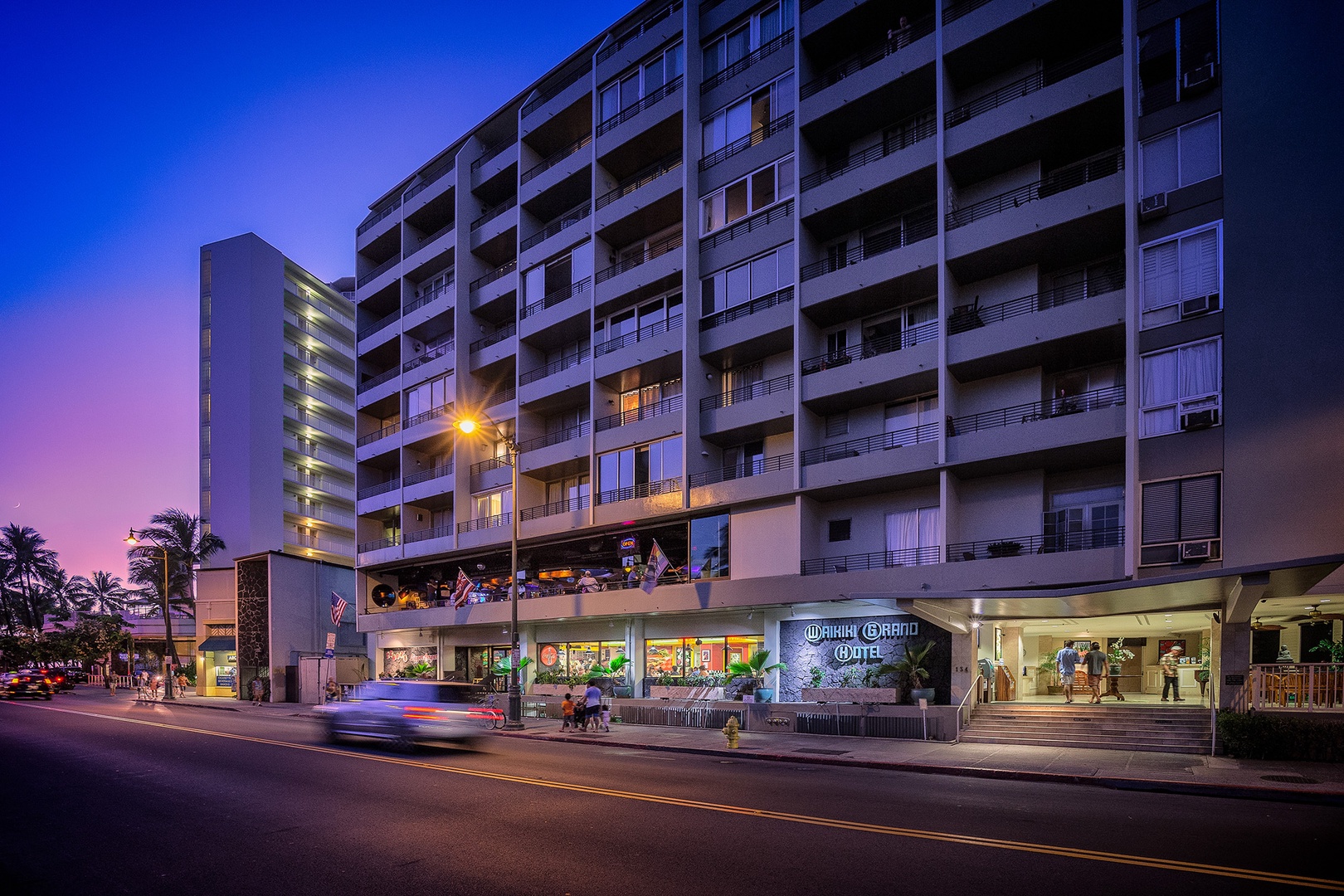 The property sits on a bustling street corner, illuminated by soft evening light with nearby shops and restaurants visible along the ground floor. A vibrant sunset paints the sky above.