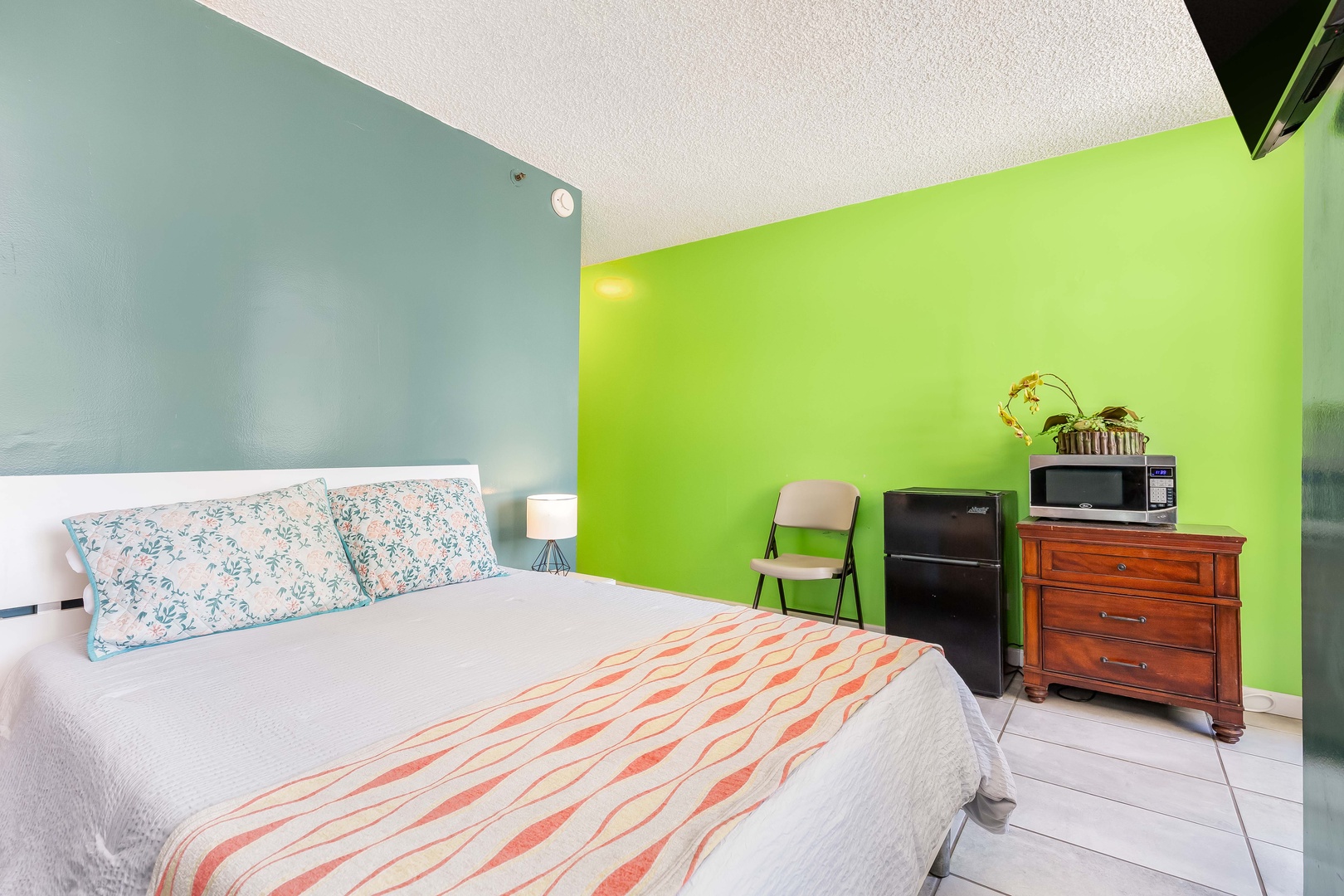 Your cozy bedroom features a comfy bed dressed in cheerful linens, pops of tropical color, and everything you need—mini fridge, microwave, and workspace—for a relaxing stay.