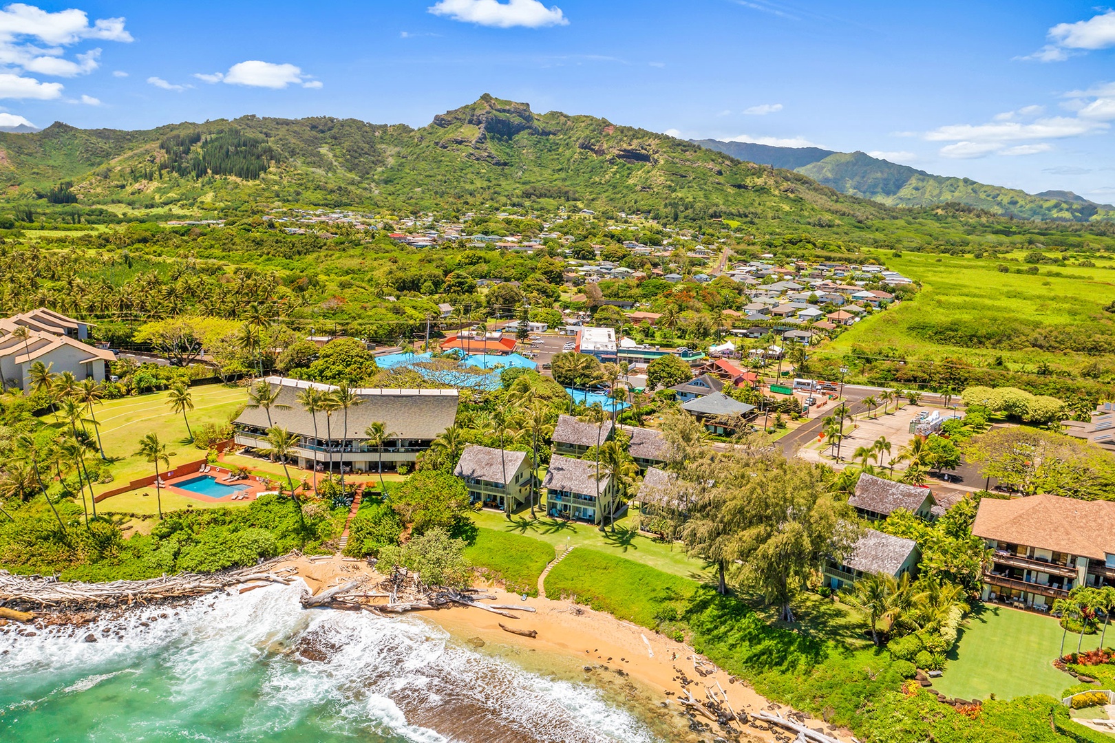 Aerial view of tropical beachfront resort nestled between turquoise waters and lush green mountains in a picturesque Hawaiian setting.