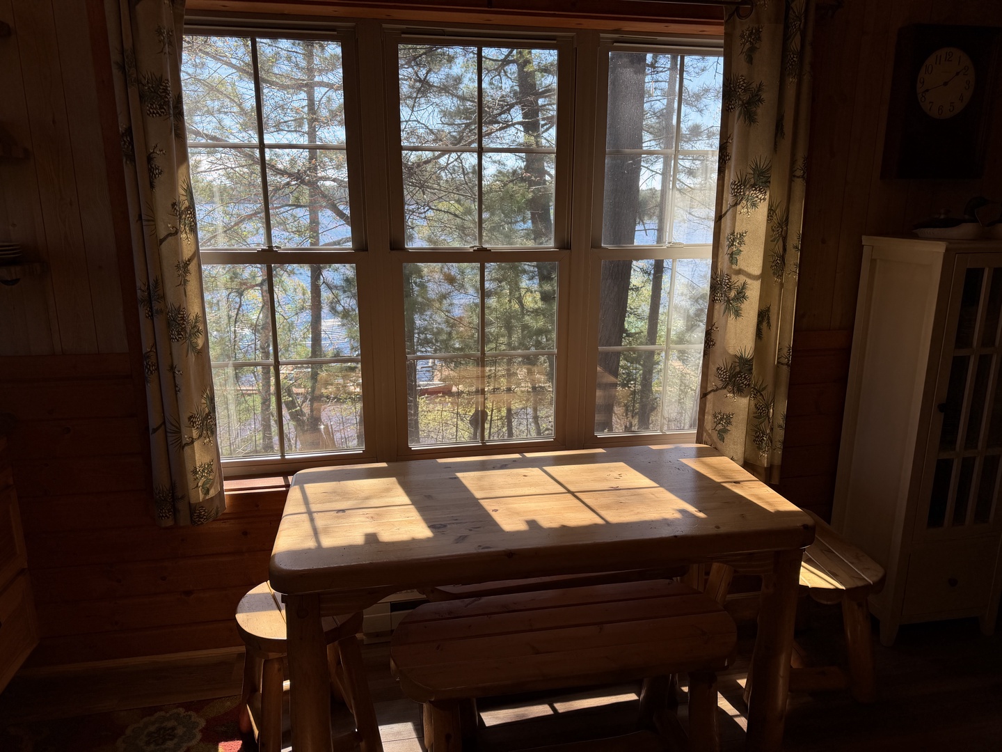 Gather around this charming wooden dining table as natural light streams through tall windows, framing peaceful lake views and tall pines—your perfect spot for morning coffee or family meals.