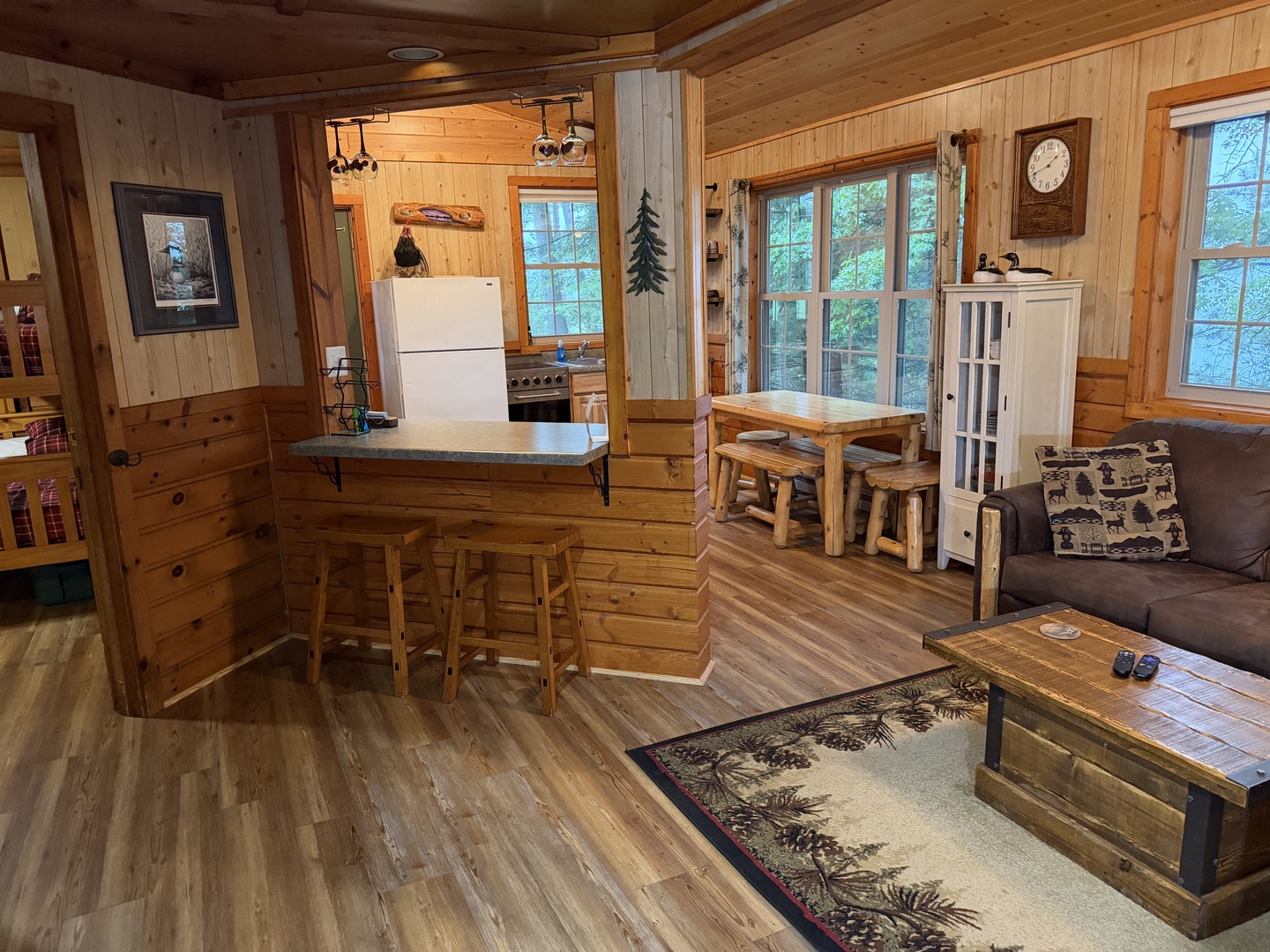 Relax in your cozy, open-concept cabin where rustic log furniture and warm wood paneling create a true mountain retreat. Sunlight streams through windows overlooking lush greenery, inviting you to unwind.