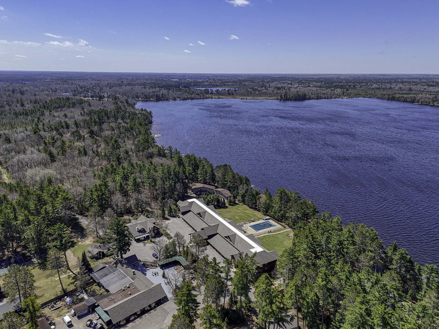 Aerial view of a lakefront property surrounded by dense forest with multiple buildings and a pool nestled along the pristine shoreline of a vast, tranquil lake.
