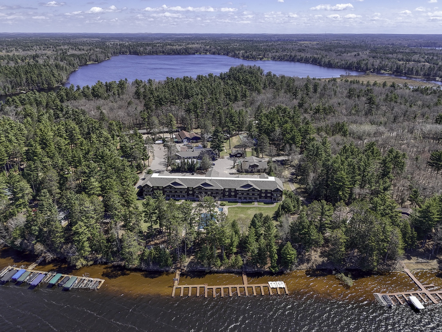 Aerial view of a lakefront vacation property nestled among towering pines, with a scenic lake and dense forest stretching to the horizon, offering a peaceful natural setting.