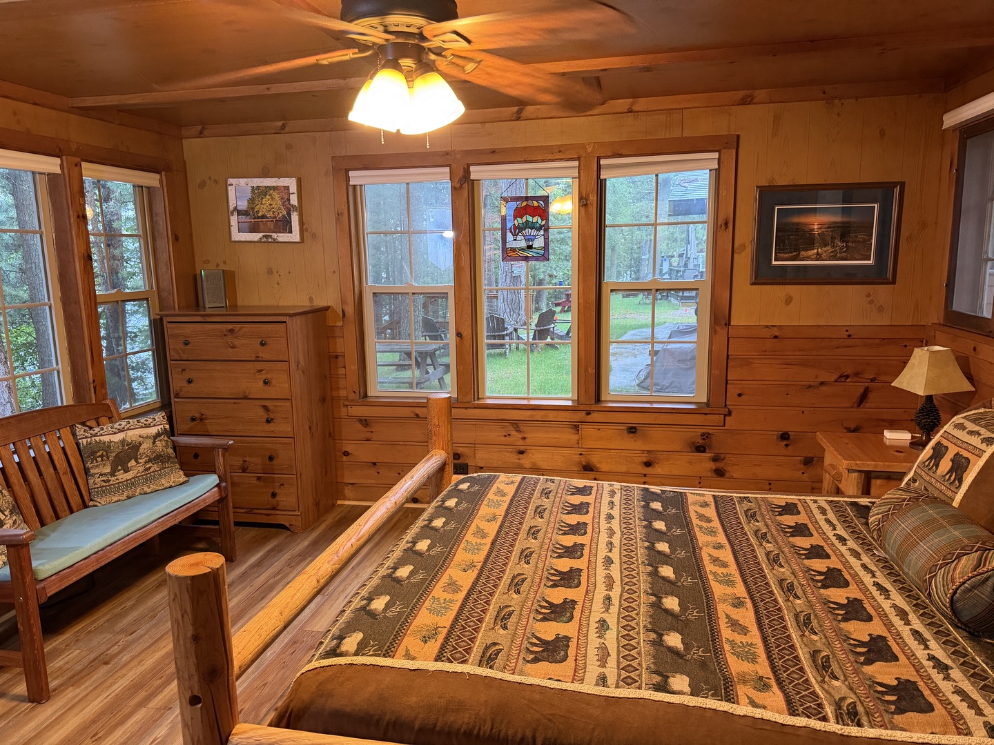 Unwind in your cozy cabin bedroom with rustic log furnishings, warm wood paneling, and peaceful garden views through generous windows that fill the space with natural light.
