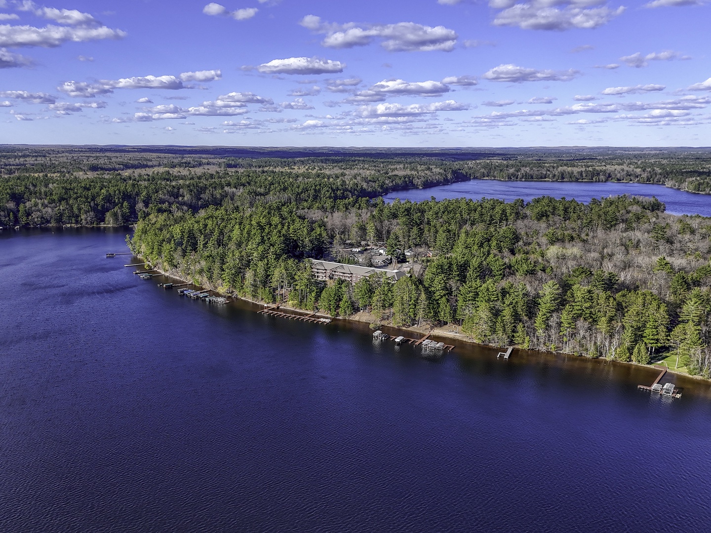 Aerial perspective of a lakefront vacation property nestled on a peninsula surrounded by dense forest, showcasing  docks and pristine waterfront access beneath a dramatic cloud-filled sky.