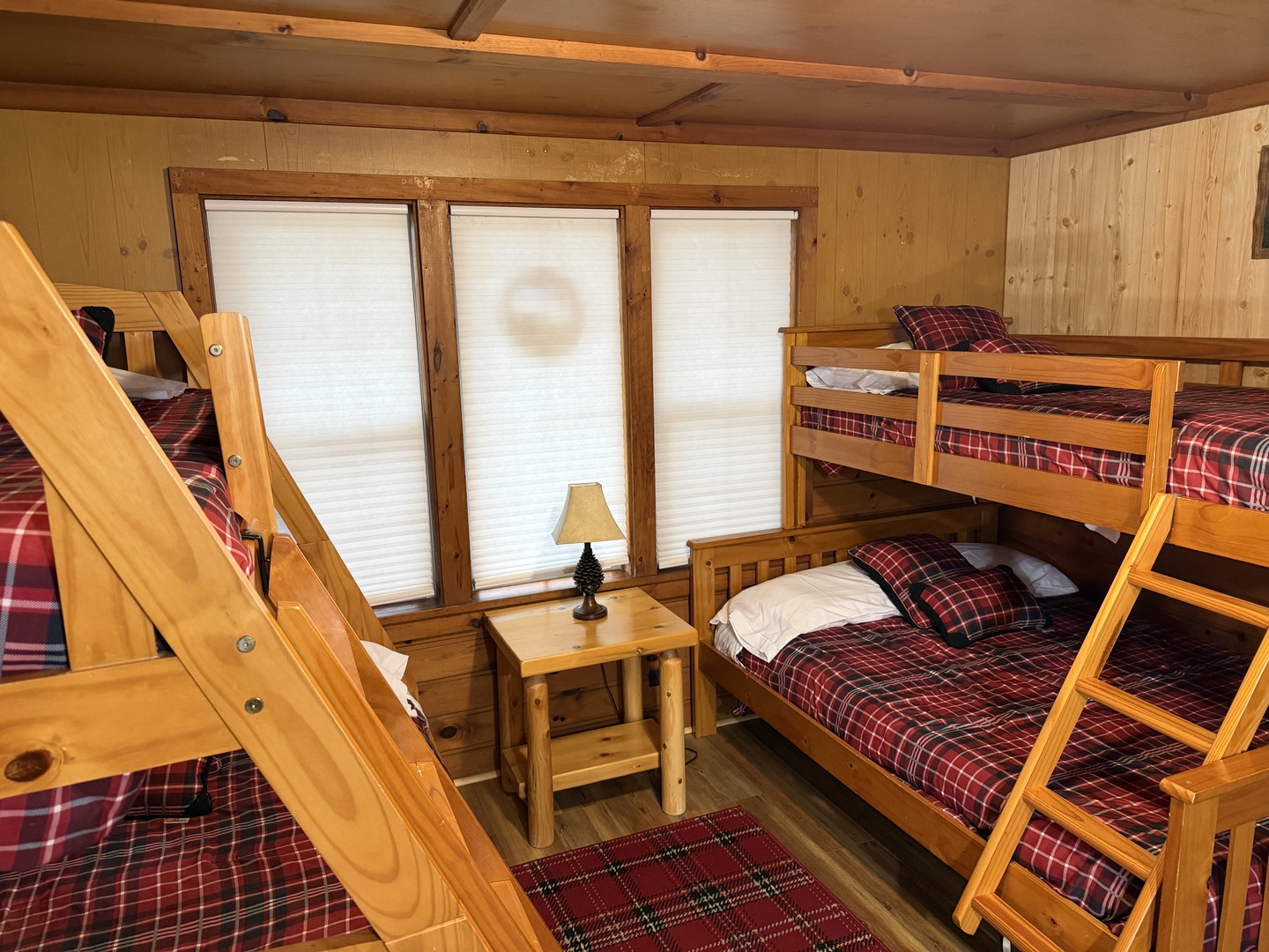 Your cozy bunkroom features warm wood paneling and plaid bedding, perfect for families or groups. Natural light filters through the windows, creating a welcoming mountain retreat atmosphere.