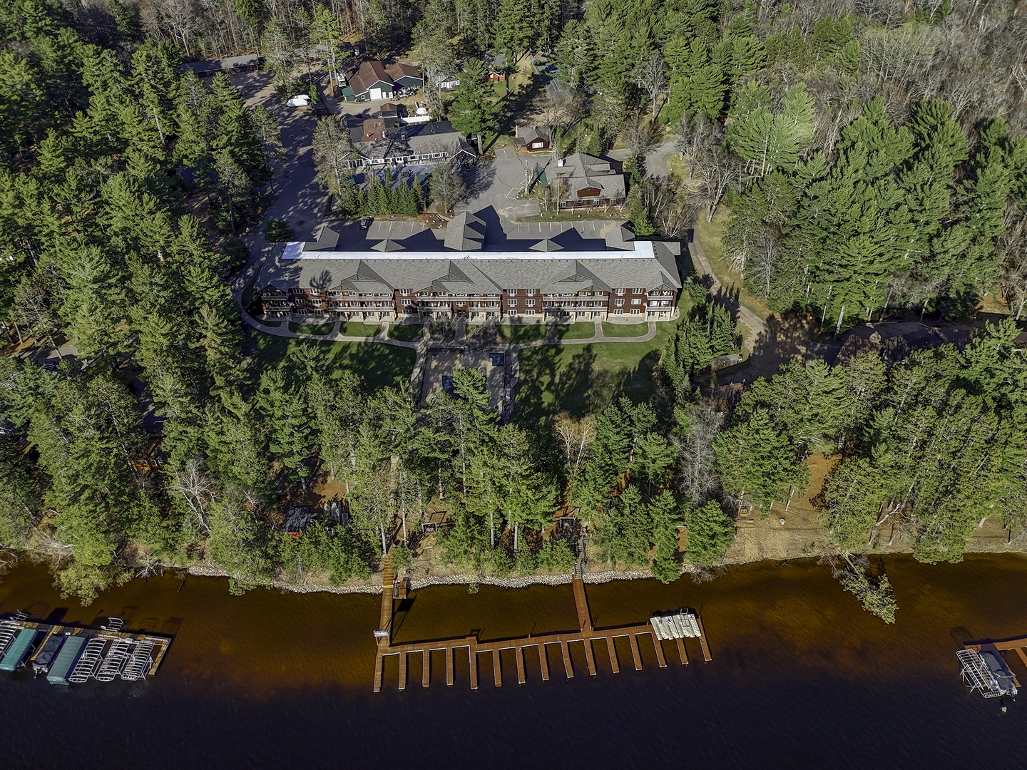 Aerial view of a lakefront property nestled among evergreen forests, featuring a main lodge building with  dock access and boat slips on the waterfront.