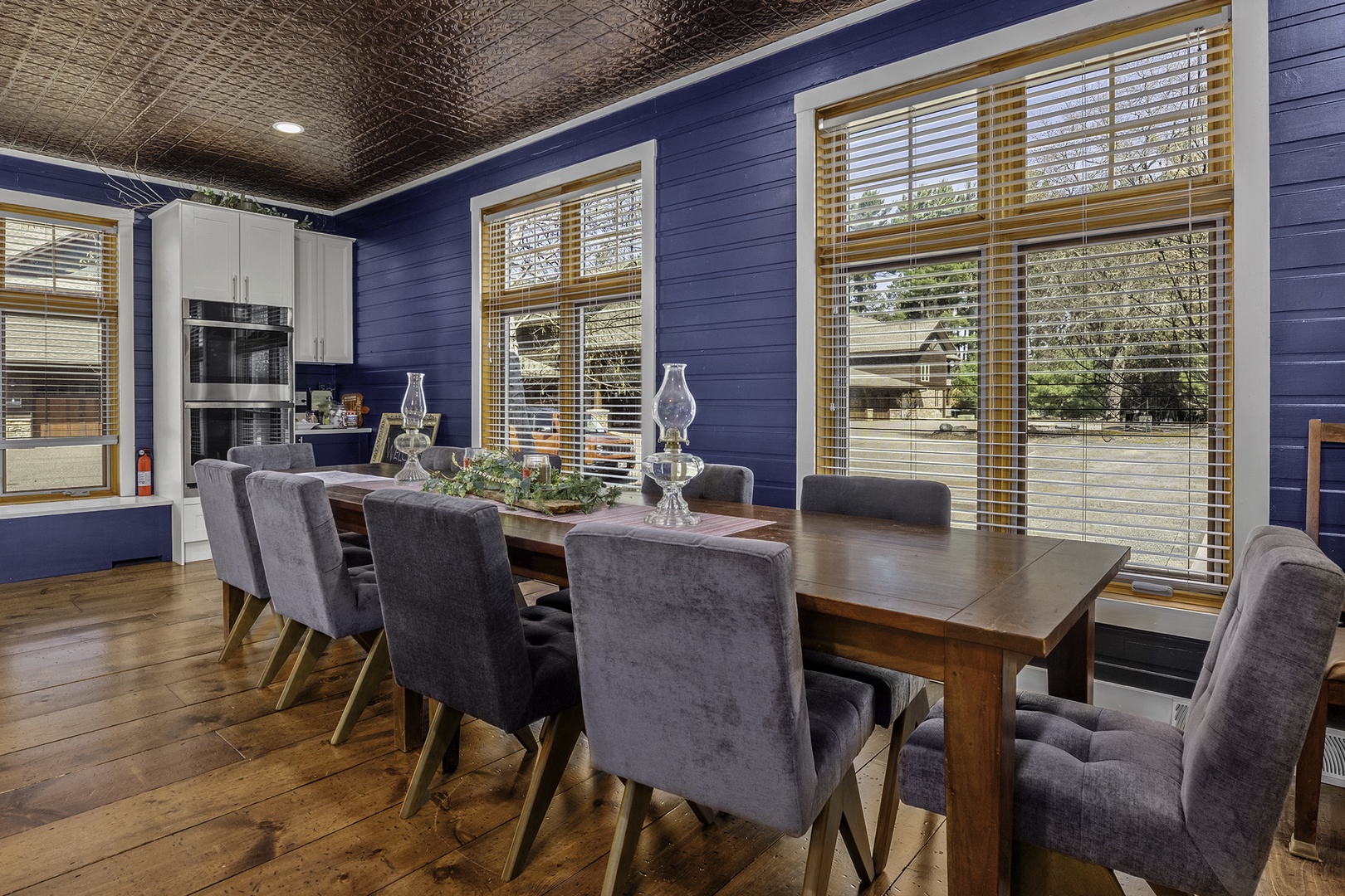 Gather around this elegant dining table where rich wood tones meet plush seating and natural light streams through charming windows, creating your perfect backdrop for memorable meals and conversation.
