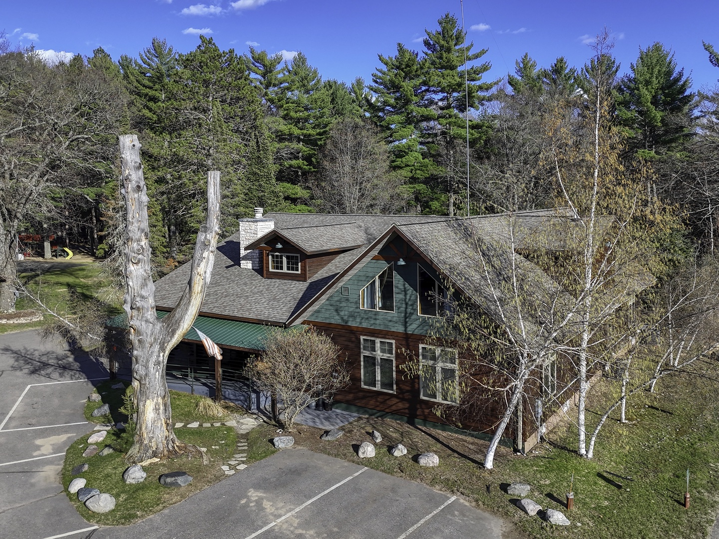 An aerial view captures the property's charming cabin-style architecture nestled among towering evergreens and birch trees, with convenient parking available at the entrance.