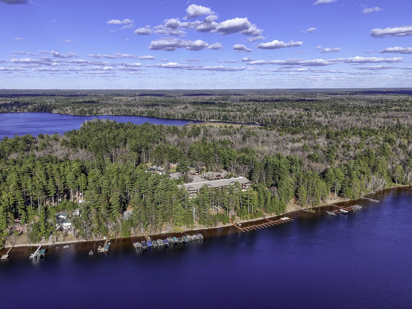 Aerial view of a lakeside vacation property nestled among dense pine forests with private docks and boat slips along pristine blue waters stretching to the horizon.