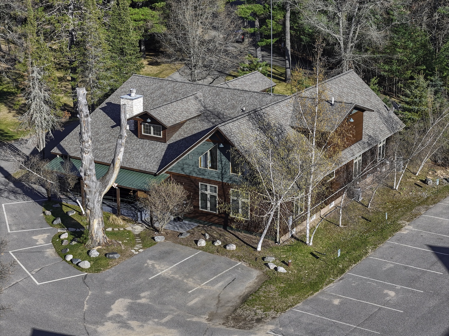 A spacious  lodge featuring rustic wood and stone architecture, nestled among tall pines and birch trees with convenient parking at the entrance.
