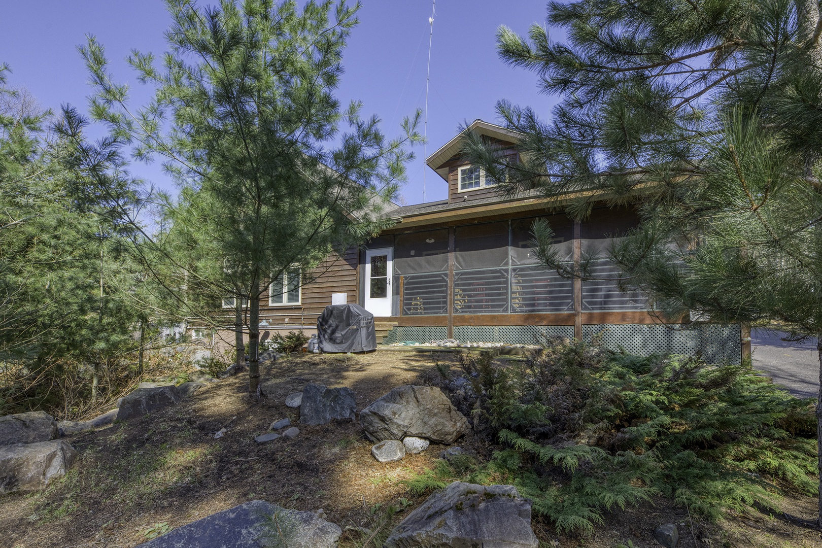Charming cabin-style property nestled among evergreens and natural rock landscaping, offering a peaceful retreat setting in a wooded area.