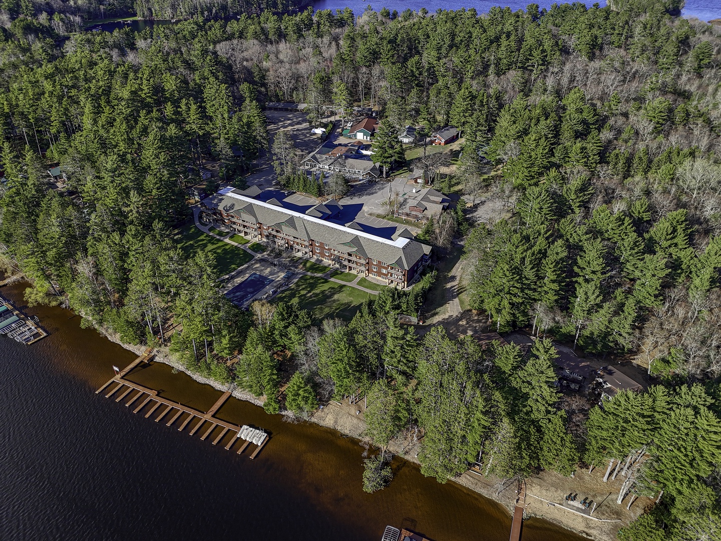 Aerial view of a secluded waterfront property nestled among towering pines, featuring multiple buildings,  dock access, and expansive natural surroundings for a peaceful retreat.