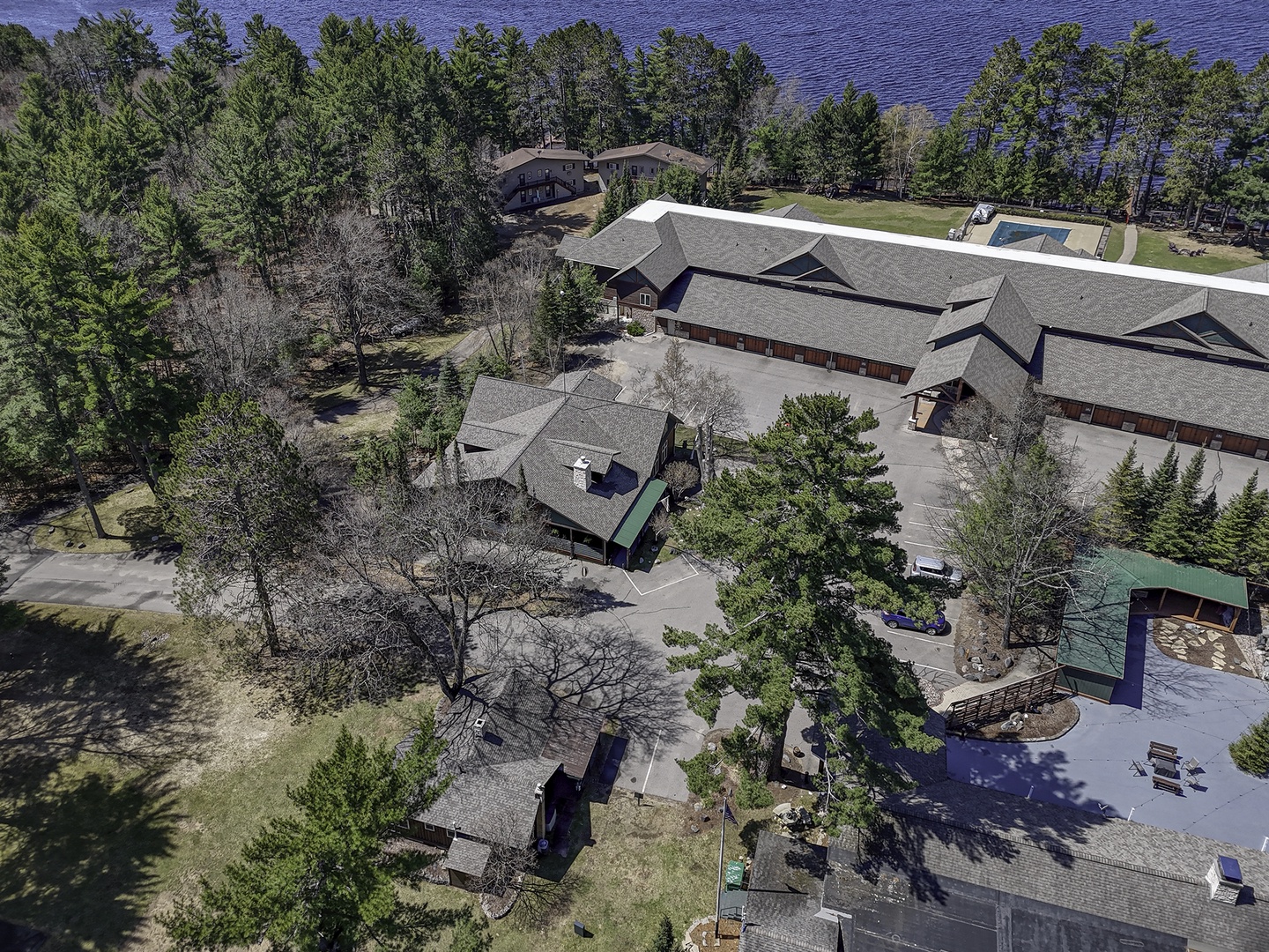 Aerial view of the property showcasing lodge-style buildings surrounded by pine forest with lake views visible in the background.