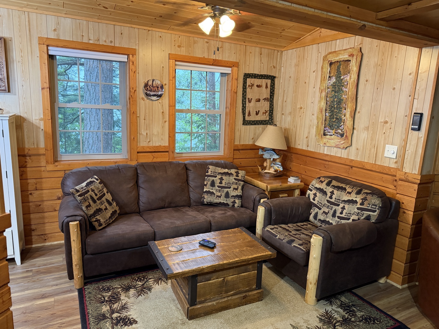 Sink into your cozy cabin living room surrounded by warm pine walls and rustic decor. Natural light streams through large windows overlooking peaceful woodlands as you unwind on plush seating.