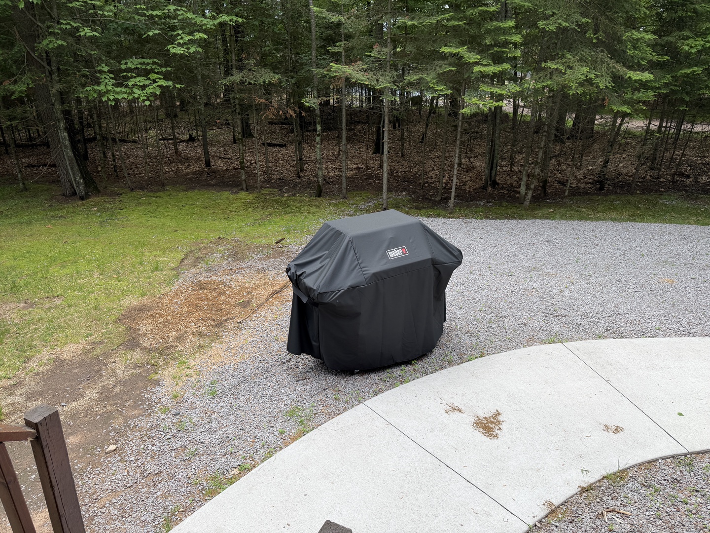 A covered grill sits ready on the gravel driveway, surrounded by peaceful woodlands that frame the property's natural setting.
