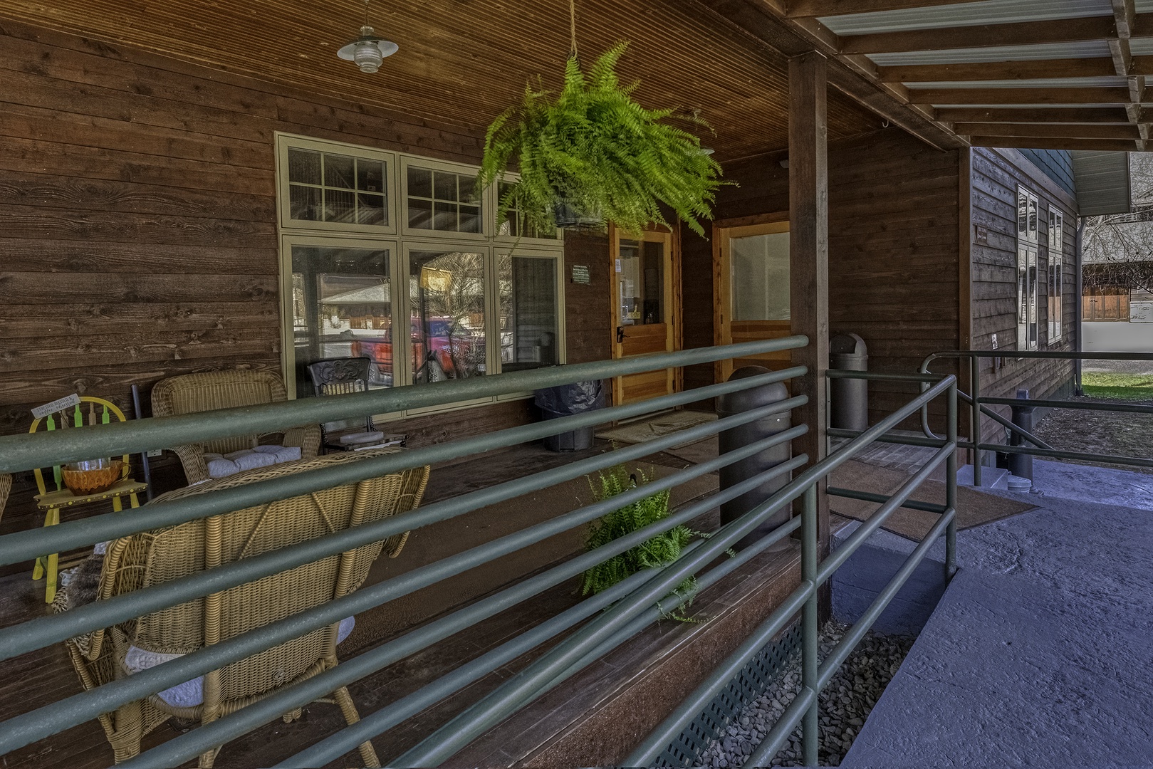 Covered porch entry features rustic wooden walls, accessible ramp with metal railings, and charming fern planters welcoming guests to the property entrance.